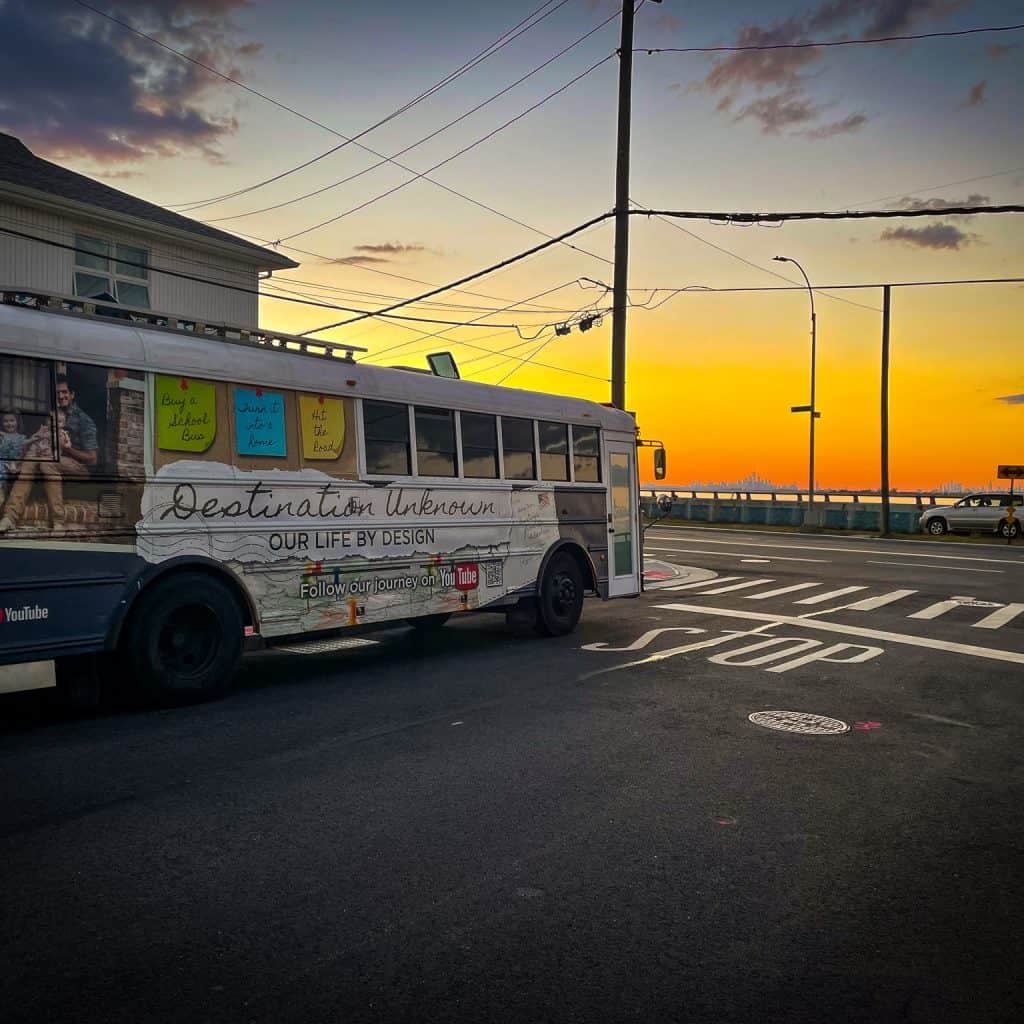 School bus tiny home sitting on the streets in Rockaway, New York, overlooking the channel to downtown Manhattan at sunset. The sky is orange and yellow. | What is a skoolie roof raise? | Destination Unknown