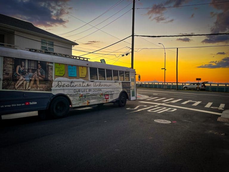 School bus tiny home sitting on the streets in Rockaway, New York, overlooking the channel to downtown Manhattan at sunset. The sky is orange and yellow. | What is a skoolie roof raise? | Destination Unknown