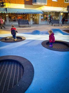 Two toddlers jumping on the in-ground trampoline in downtown Sandviken, Sweden. The month is October so the children are wearing winter coats because of the cold. | Destination Unknown