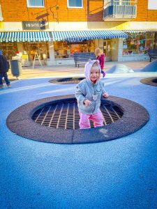 Young girl jumping on in-ground trampoline in downtown Sandviken, Sweden. The girl is wearing a gray wool jacket with pink jumper underneath. She has light blonde hair. | Destination Unknown