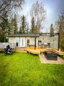 A small green Swedish cottage called Högbro Brukstuga. The cottage is surrounded by a wooden wrap around porch with table, chairs and firepit. Behind the cottage are tall pine and birch trees showing fall colors. | Sandviken Sweden | Destination Unknown