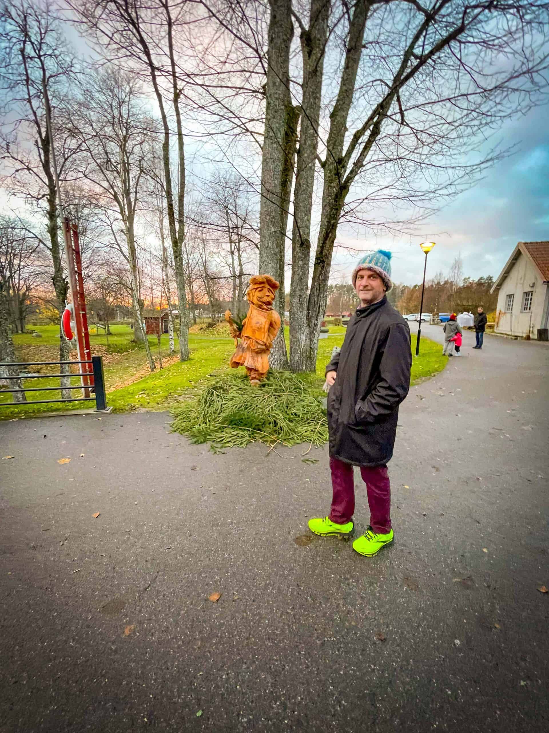 Man dressed in warm clothes and neon yellow sneakers standing next to a wooden statue of the Swedish Chef at Höbro Bruk in Sandviken, Sweden | Destination Unknown