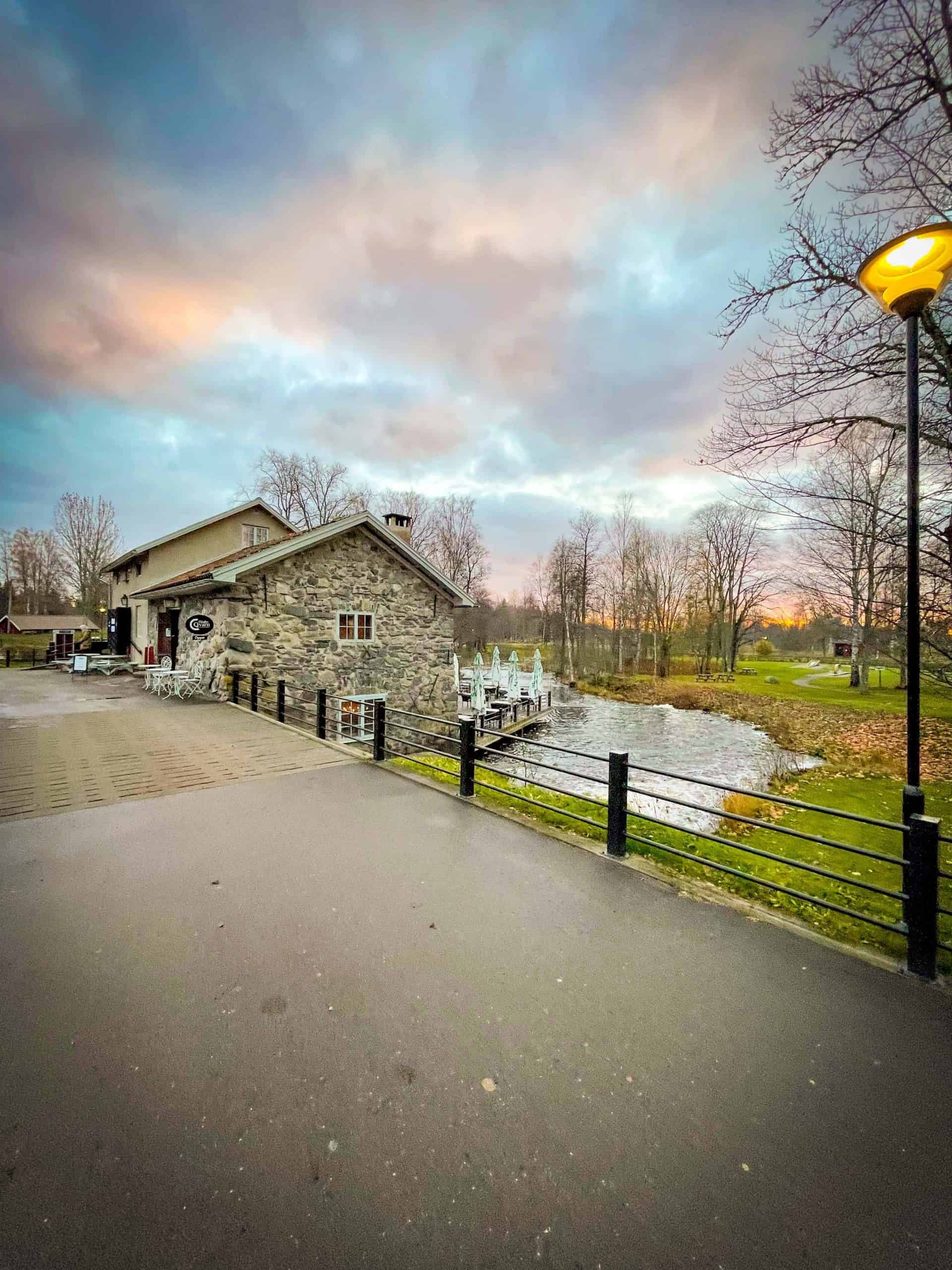 A small bridge going over the river towards a gray stone Swedish cottage in at the Höbro Bruk in Sandviken, Sweden. The sun is setting and the sky looks orange, blue and pink. There are no leaves on the trees in October. | Destination Unknown