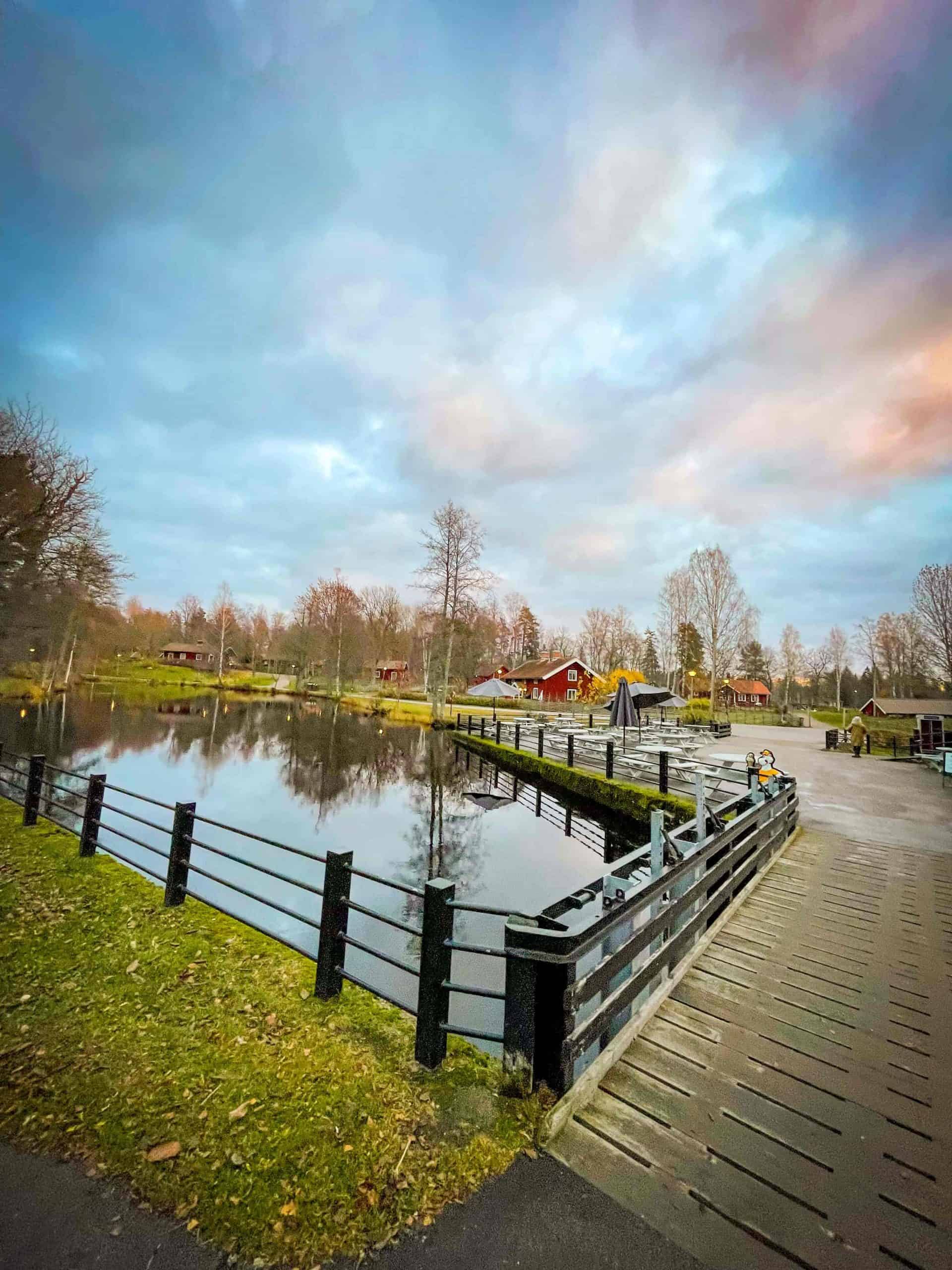 A small bridge going over the river in the foreground. In the background are red Swedish cottages at the Höbro Bruk in Sandviken, Sweden | Destination Unknown