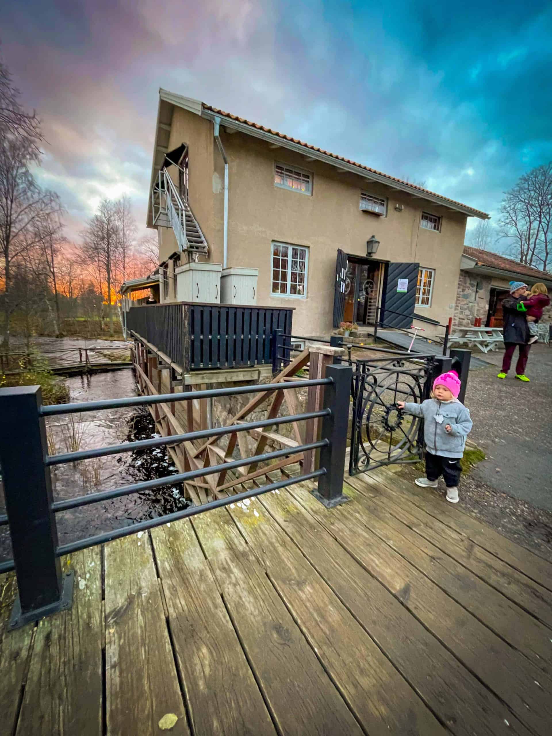Young girl wearing a pink hat and gray wool sweater, holding the railing of a bridge by the restaurant at Höbro Bruk in Sandviken, Sweden | Destination Unknown