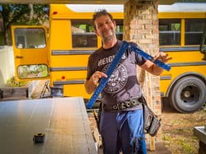 Man holding a t-square in foreground. A yellow school bus conversion is behind him. | what is a skoolie roof raise? | DESTINATION UNKNOWN