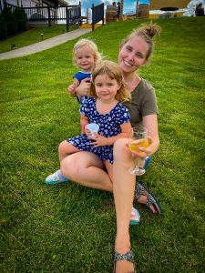 Mom and two daughters smiling and sitting in the grass. Mom has a glass of white wine in her hand.