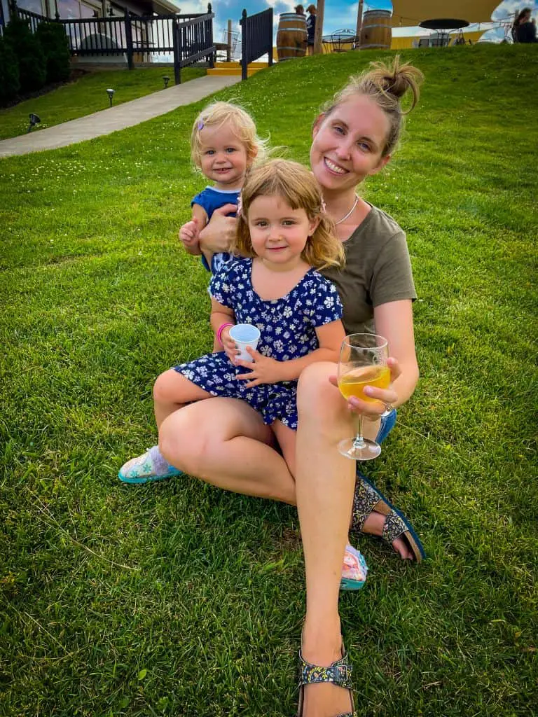 Mom and two daughters smiling and sitting in the grass. Mom has a glass of white wine in her hand.