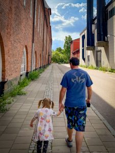 Father and daughter walking through a historic area in Västerås Sweden. There is a red brick warehouse on the left and a factory with black smoke stacks on the right.