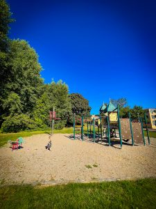 photo of a little girl playing on a Swedish playground. The sky is bright blue. There are pebbles in the play area. The playground has trees on the left.
