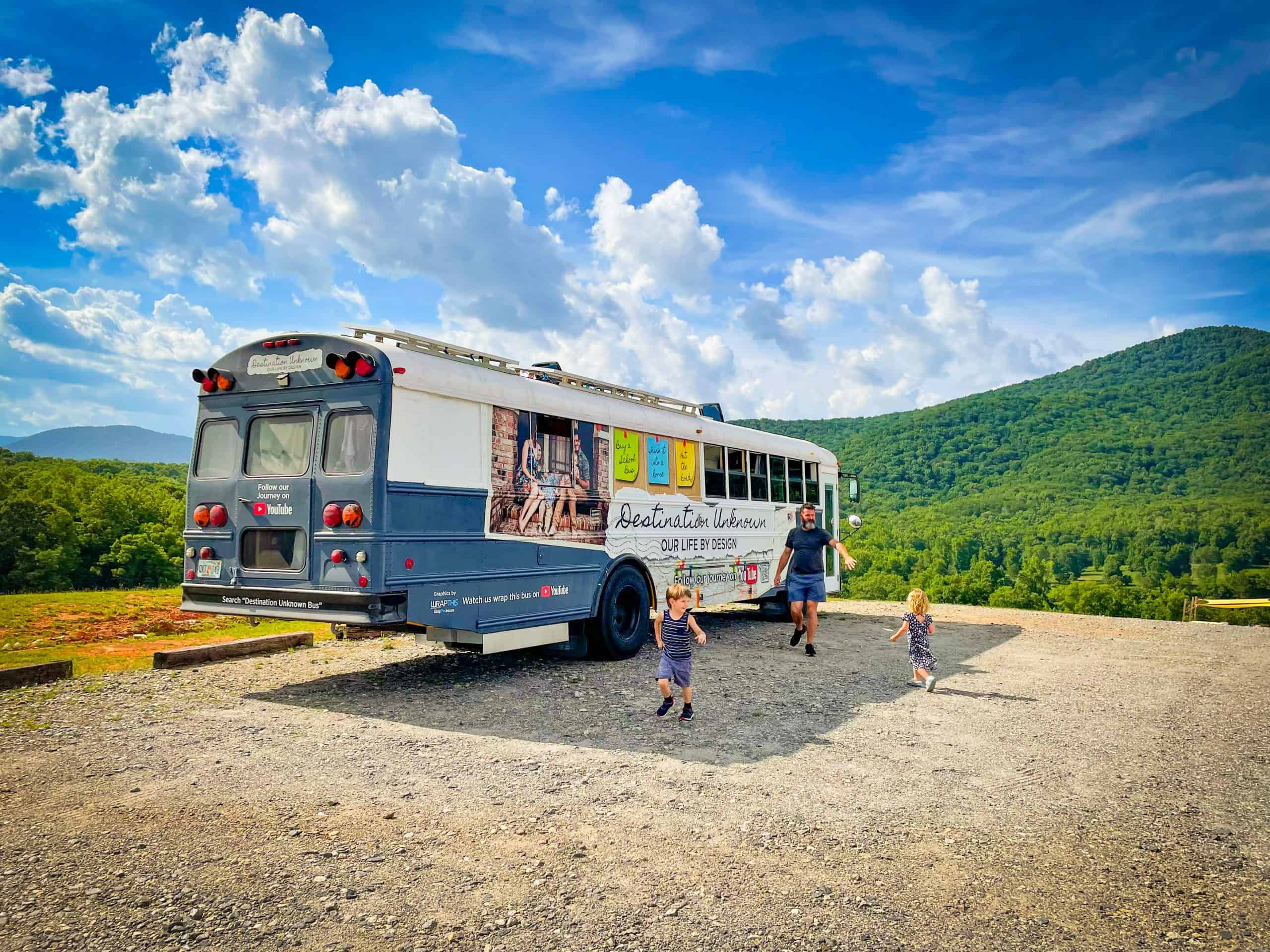 Blue and white school bus conversion RV parked on a mountain. A father with two children are running at the camera.